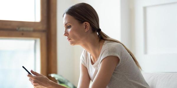 Side view thoughtful young woman sitting on couch, looking at mobile phone screen, waiting for message from beloved man, feeling lonely or jealous. Unhappy lady reading news in social media.