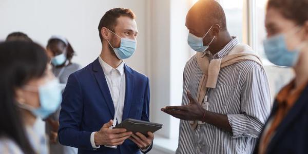 Waist up portrait of group of people chatting during break at business conference, focus on two men wearing masks with lens flare